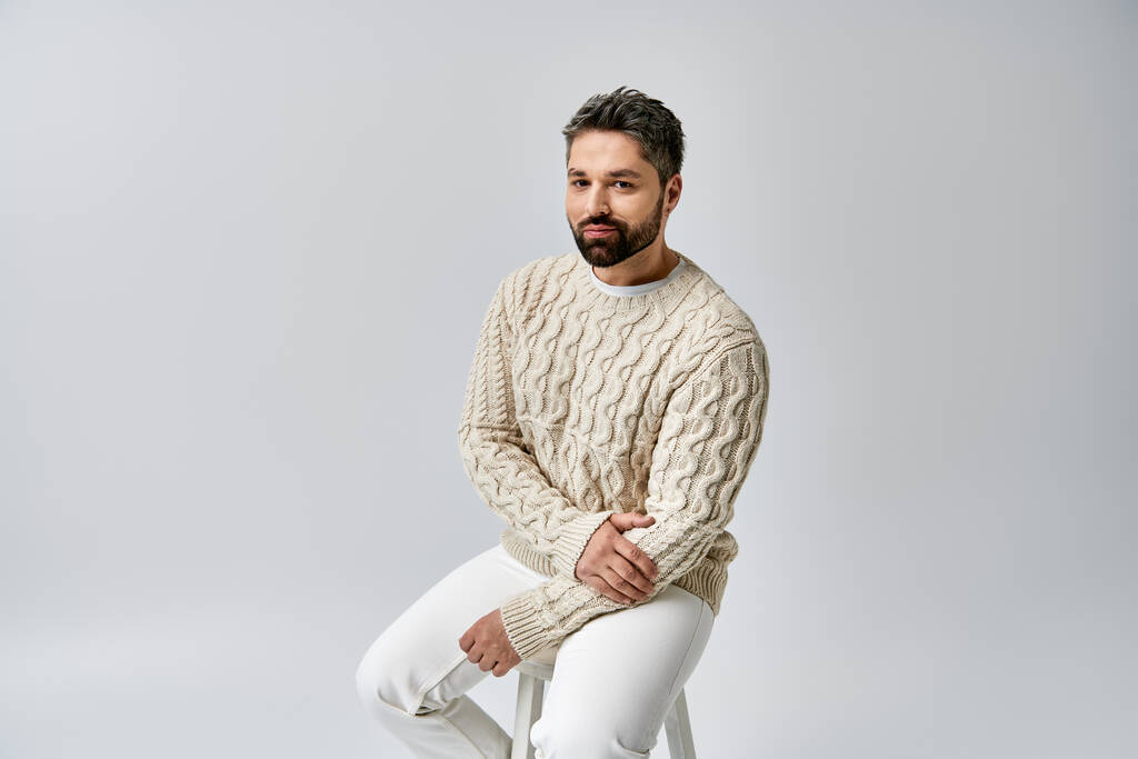 Un hombre carismático con barba se sienta en un taburete, exudando encanto en un acogedor suéter blanco sobre un fondo gris del estudio. - Foto, Imagen