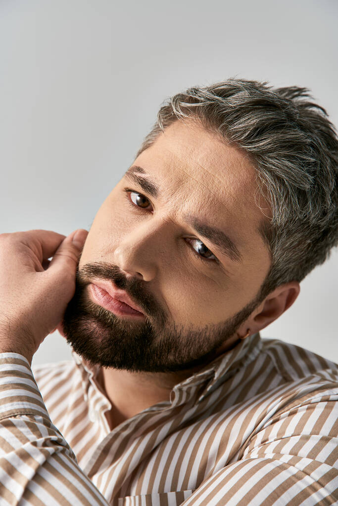 Un hombre con estilo con una barba exudando encanto en una camisa a rayas sobre un fondo de estudio neutral. - Foto, imagen