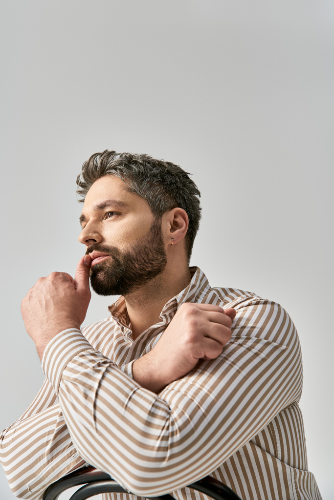 Un hombre sofisticado con barba posa con confianza en una camisa a rayas sobre un fondo gris del estudio. - Foto, Imagen