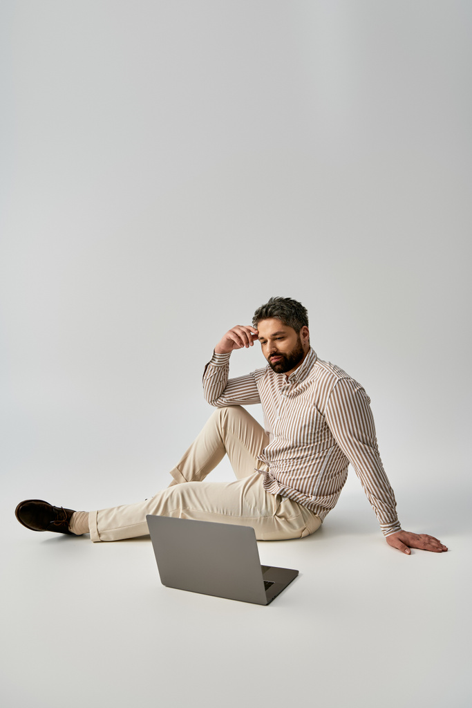 Un uomo alla moda con la barba si siede a terra, indossando abiti eleganti, mentre lavora intensamente sul suo computer portatile.. - Foto, immagini
