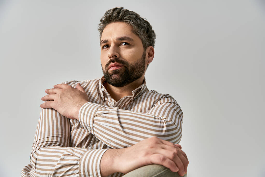Un homme élégant avec une barbe frappant une pose dans une chemise rayée sur fond de studio gris. - Photo, image