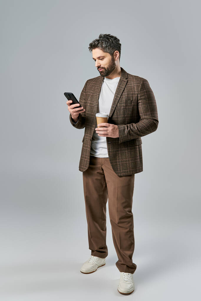 Un hombre elegante con un atuendo elegante sosteniendo una taza de café y revisando su teléfono mientras se pierde en el pensamiento. - Foto, Imagen