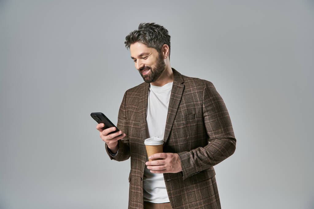 Egy kifinomult, szakállas, elegáns öltözetű férfi tart egy csésze kávét, miközben nézi a mobilját.. - Fotó, kép