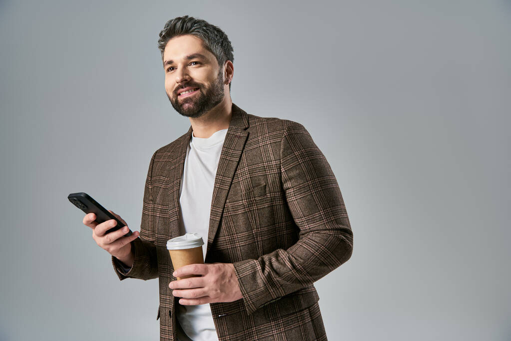 Un uomo barbuto in abbigliamento elegante tiene una tazza di caffè mentre controlla il suo telefono cellulare, trasudando raffinatezza e multitasking. - Foto, immagini