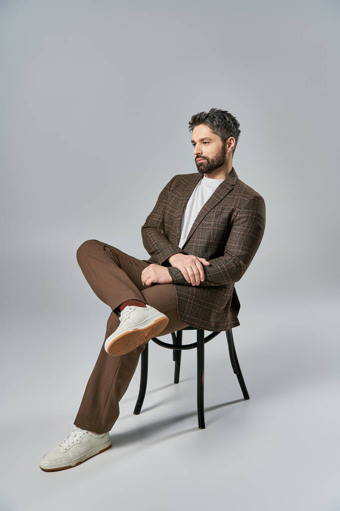 Důmyslný muž s vousy sedí elegantně na židli, zkříží nohy, ve studiu s šedým pozadím. - Fotografie, Obrázek