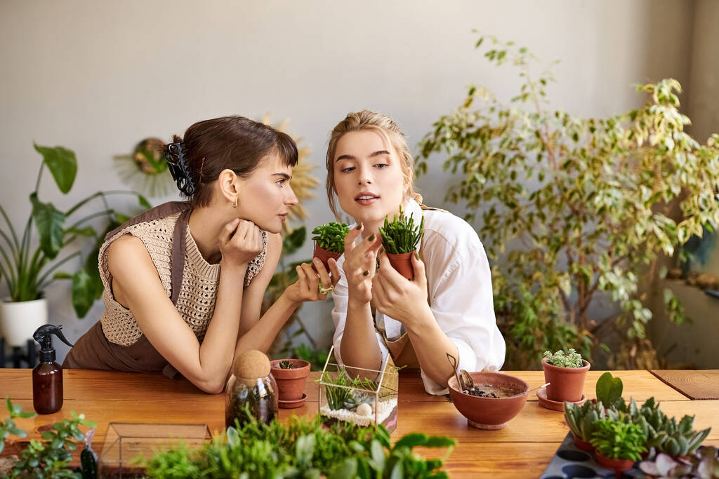 Embracing artistry, twee vrouwen zitten aan een tafel omgeven door weelderige groene planten. - Foto, afbeelding
