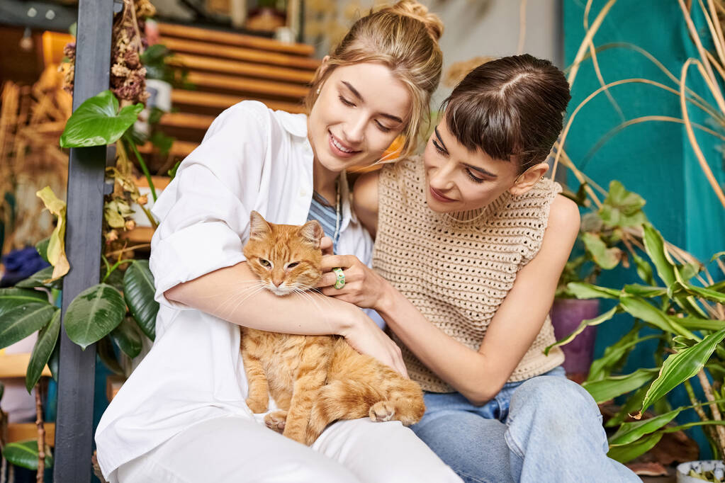 Dos mujeres, una pareja lesbiana cariñosa, se sientan tranquilamente en un porche con un gato, rodeado de decoración artística. - Foto, Imagen