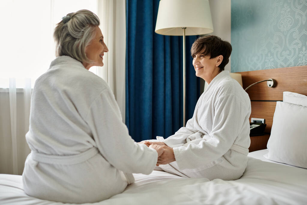 Senior lesbian couple sitting together on a bed, wearing robes, sharing a tender moment. - Photo, Image