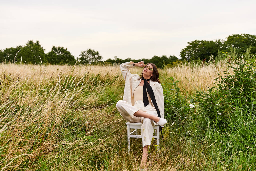 Молода жінка, одягнена в білий колір, мирно сидить у кріслі серед пишного поля, заспокоюючись в теплі літнього бризу. - Фото, зображення