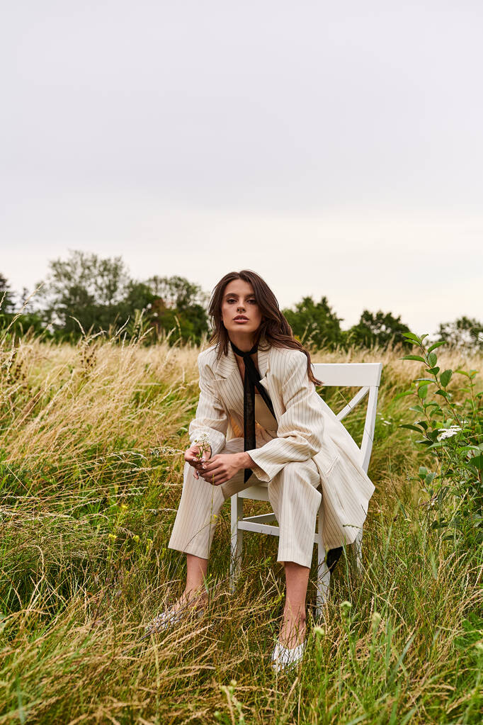 Μια όμορφη νεαρή γυναίκα με άσπρη ενδυμασία κάθεται σε μια καρέκλα σε ένα ήσυχο χωράφι, μουλιάζοντας στο καλοκαιρινό αεράκι. - Φωτογραφία, εικόνα