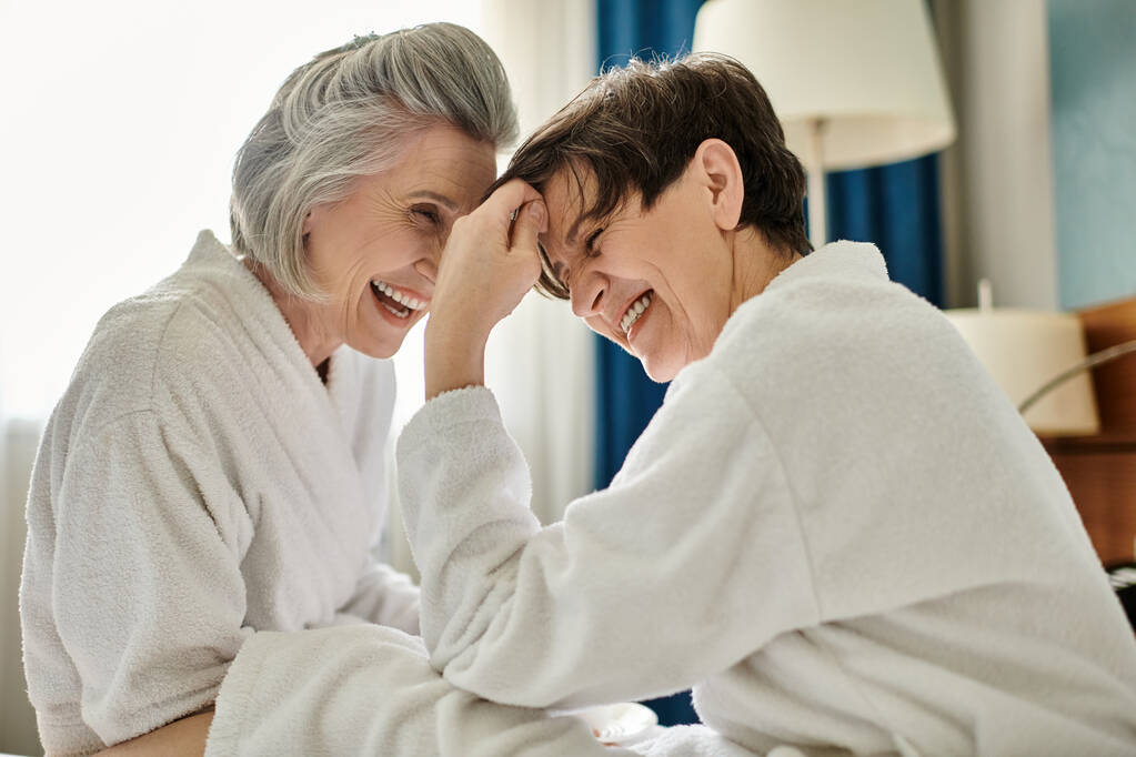 2人の上級女性が居心地の良いベッドで笑いの瞬間を共有する. - 写真・画像