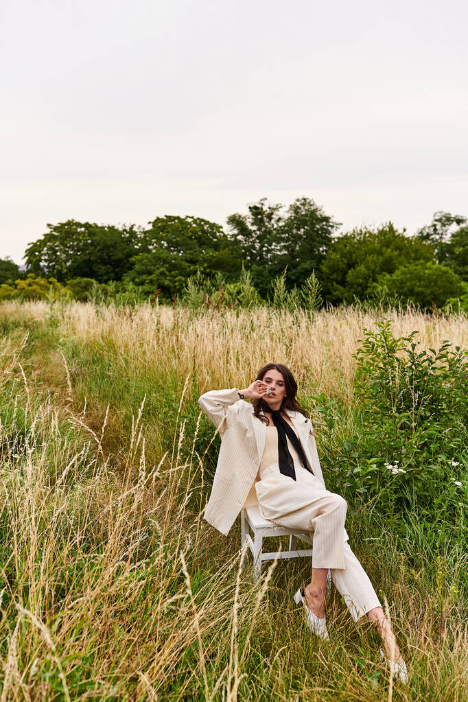 Uma bela jovem mulher de traje branco senta-se em uma cadeira em um campo beijado pelo sol, desfrutando serenamente da brisa do verão.. - Foto, Imagem