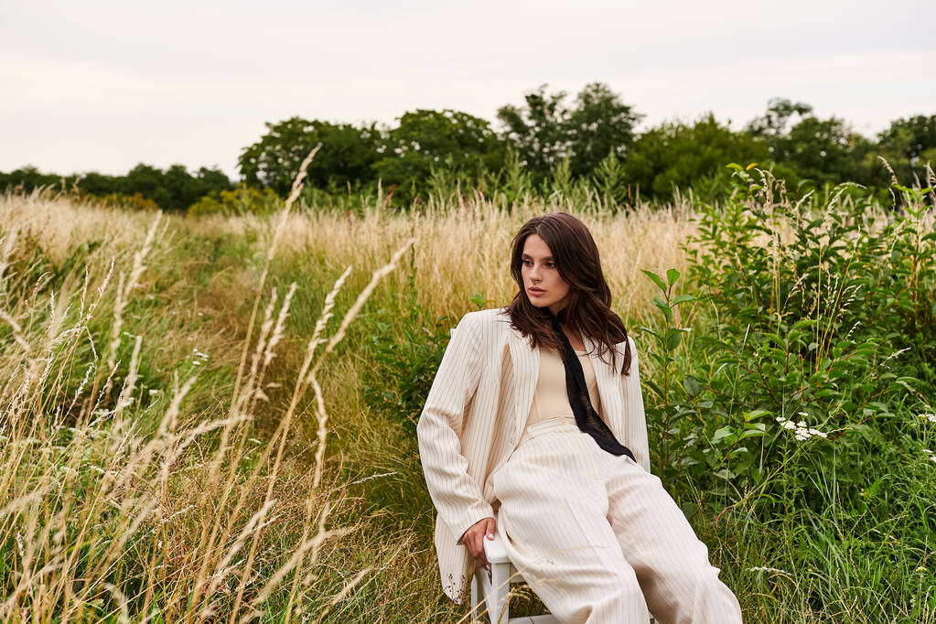 Een mooie jonge vrouw in witte kledij zit rustig op een stoel in een veld, te genieten van de zomerbries. - Foto, afbeelding