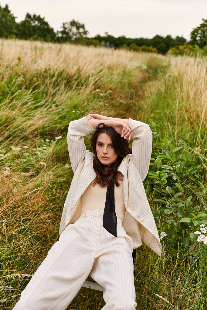 Una hermosa joven vestida de blanco sentada en un campo, con las manos en la cabeza, acariciando la brisa del verano. - Foto, Imagen