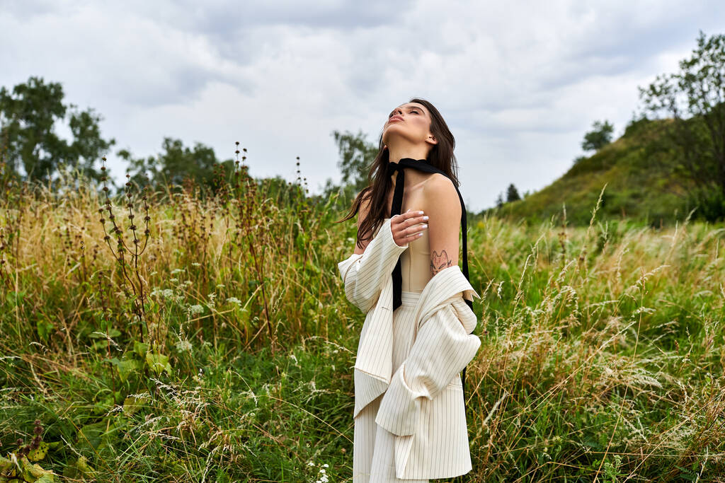 Una hermosa joven vestida de blanco de pie en un campo con los ojos cerrados, empapada en el calor del sol. - Foto, Imagen