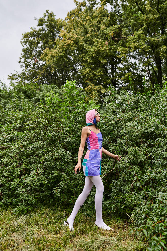 Una joven con un vestido colorido y una bufanda en la cabeza, abrazando la brisa del verano en la naturaleza. - Foto, imagen