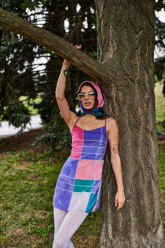 Μια όμορφη νεαρή γυναίκα με ένα ζωντανό φόρεμα και γυαλιά ηλίου στέκεται δίπλα σε ένα δέντρο, απολαμβάνοντας το καλοκαιρινό αεράκι. - Φωτογραφία, εικόνα