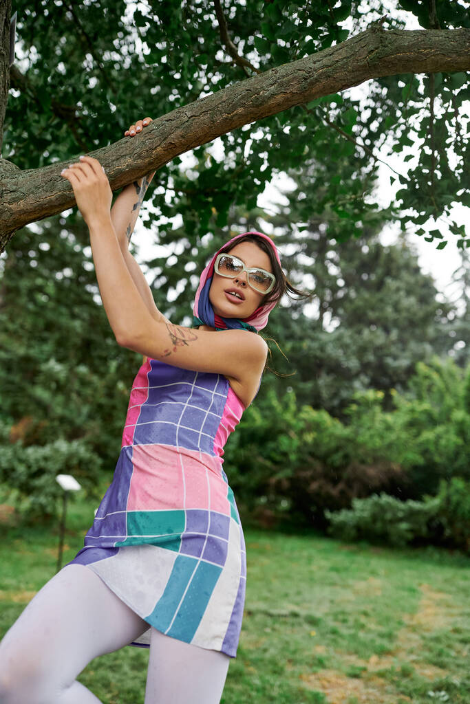 Eine schöne junge Frau in einem lebendigen Kleid und Sonnenbrille balanciert anmutig auf einem Ast und genießt die Sommerbrise. - Foto, Bild
