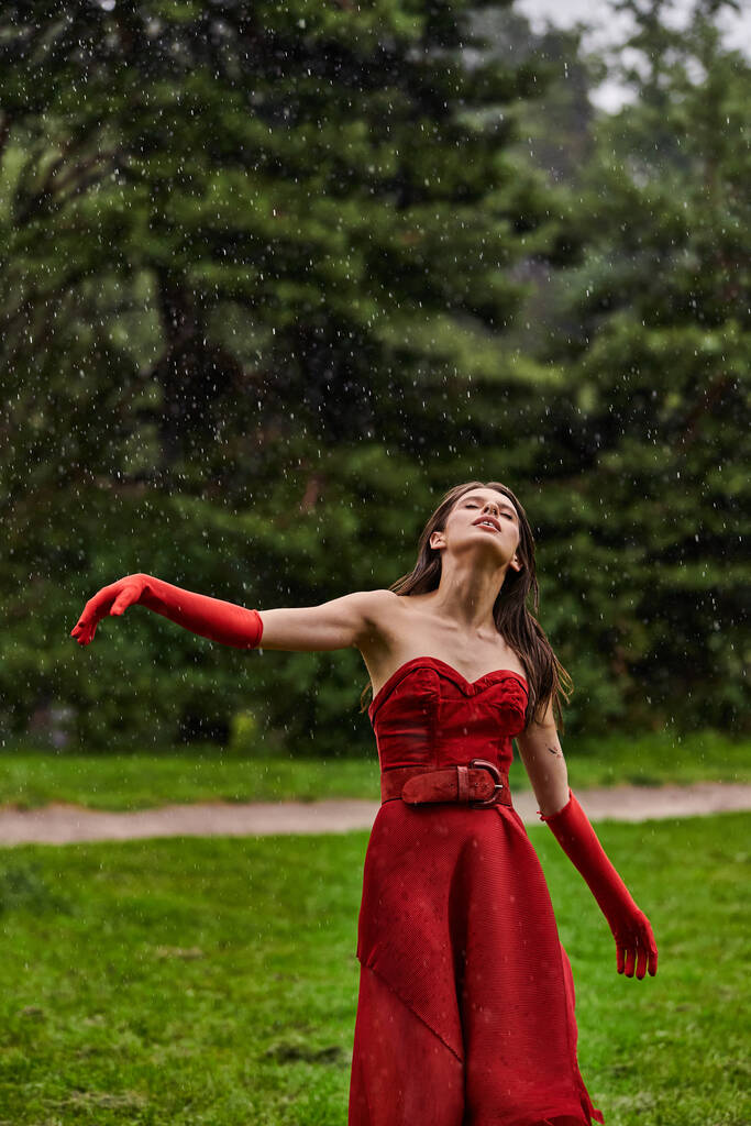 Μια ελκυστική νεαρή γυναίκα σε ένα κόκκινο φόρεμα και μακριά γάντια στέκεται με χάρη στη βροχή, απολαμβάνοντας την καλοκαιρινή νεροποντή. - Φωτογραφία, εικόνα