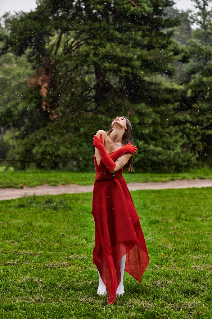 Parlak kırmızı elbiseli büyüleyici genç bir kadın zarif bir şekilde yağmurda duruyor, özgüven ve zarafetle elementleri kucaklıyor.. - Fotoğraf, Görsel