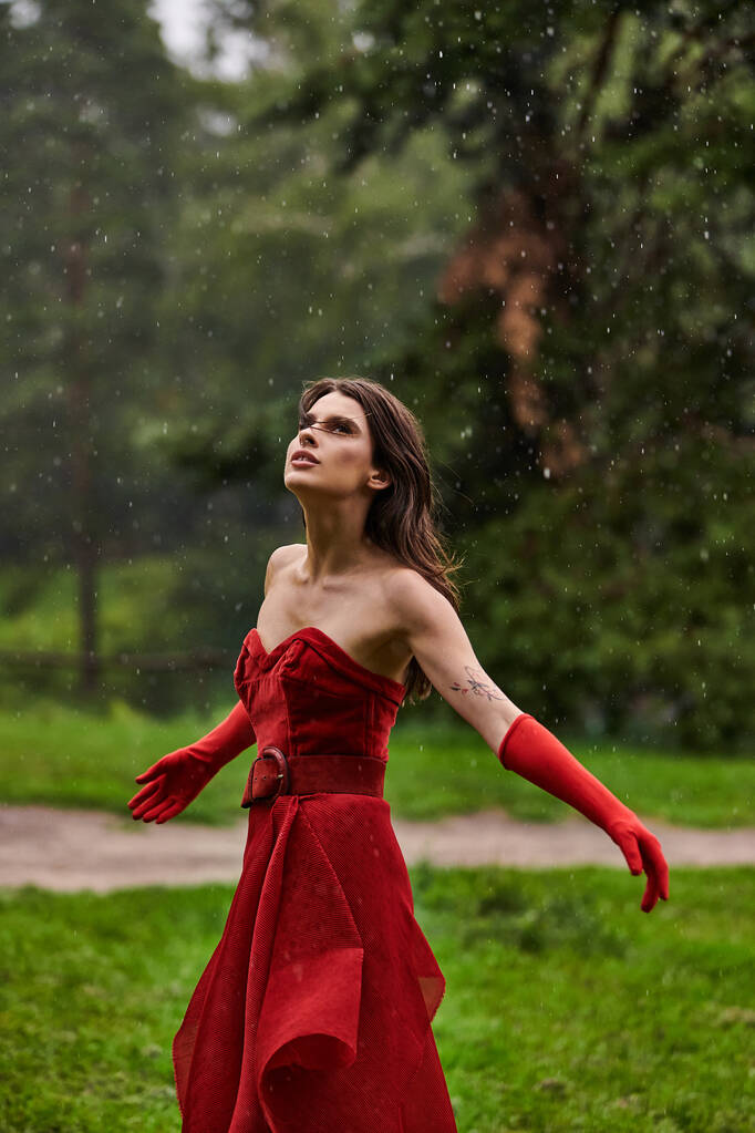 Een prachtige jonge vrouw in een opvallende rode jurk staat gracieus onder de regen, het vastleggen van de schoonheid van het moment. - Foto, afbeelding