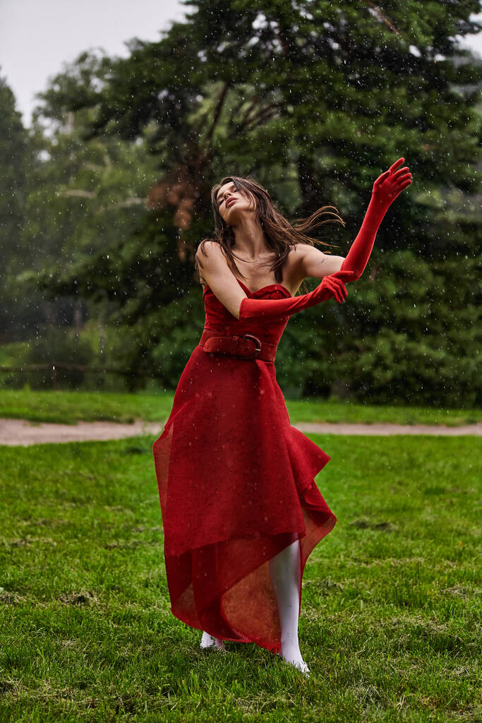 Una giovane donna in abito rosso si erge con grazia sotto la pioggia, godendo la brezza estiva in un ambiente naturale. - Foto, immagini