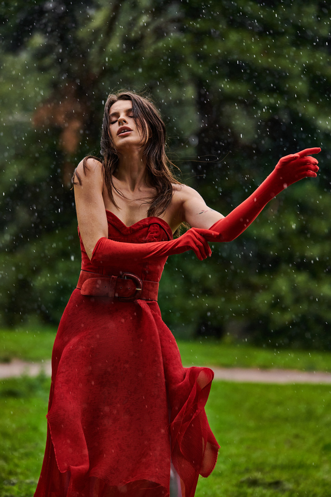 印象的な赤いドレスの若い女性は,自然の美しさを受け入れる夏の雨のシャワーで優雅に立っています. - 写真・画像