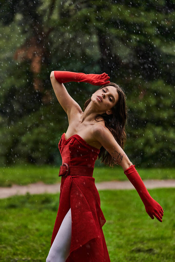 Atrakcyjna młoda kobieta w czerwonej sukience i długich rękawiczkach radośnie tańczy w deszczu, obejmując letnią bryzę. - Zdjęcie, obraz