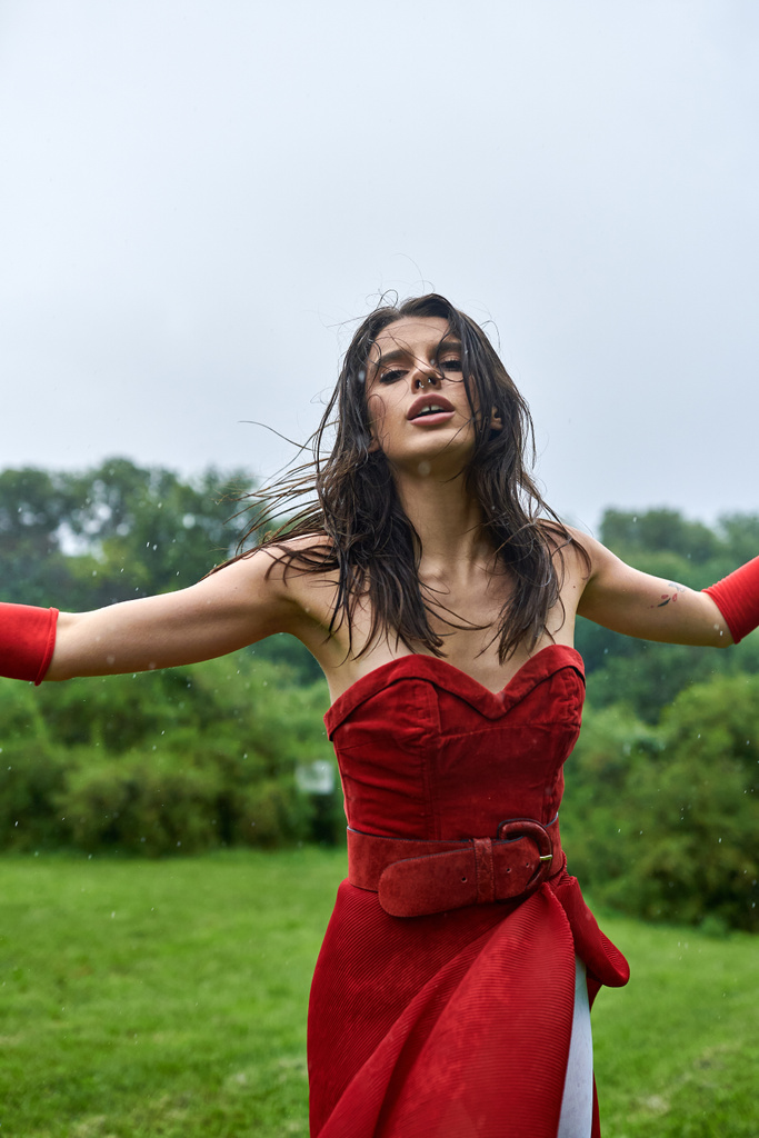 Een prachtige jonge vrouw in een rode jurk en lange handschoenen staat sierlijk in een veld, het gevoel van de zachte zomerbries. - Foto, afbeelding