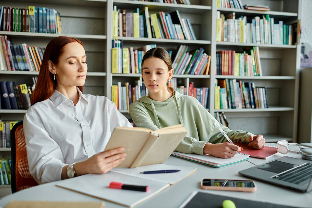 Kızıl saçlı bir öğretmen, kütüphanedeki bir masada genç bir kıza modern eğitim amaçlı dizüstü bilgisayar kullanmayı öğretiyor.. - Fotoğraf, Görsel