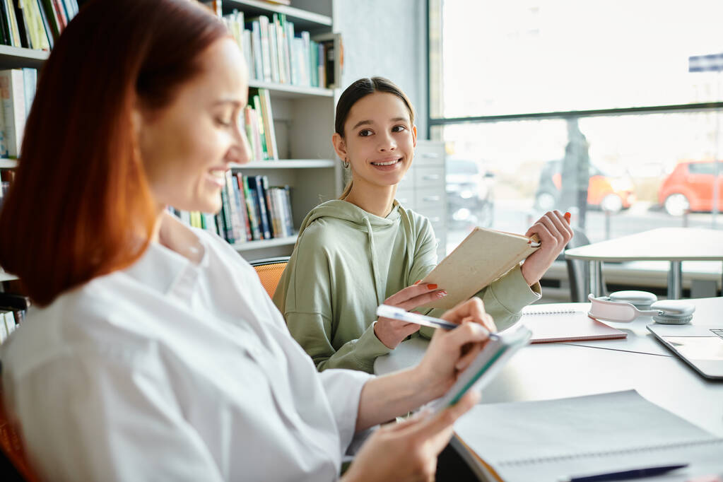 Ein Nachhilfelehrer mit roten Haaren unterrichtet ein Mädchen im Teenageralter während des Nachmittagsunterrichts, wobei er sich auf einen Laptop konzentriert, in einem modernen pädagogischen Umfeld. - Foto, Bild