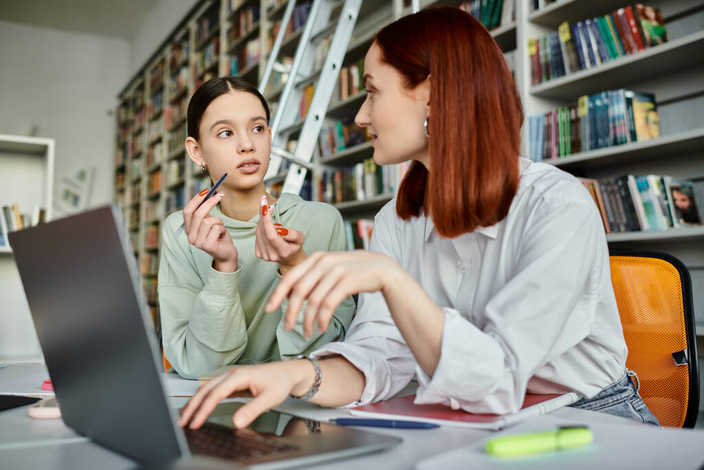 Un tuteur roux enseigne à une adolescente dans une bibliothèque, tous deux absorbés dans un ordinateur portable pendant une leçon après l'école. - Photo, image