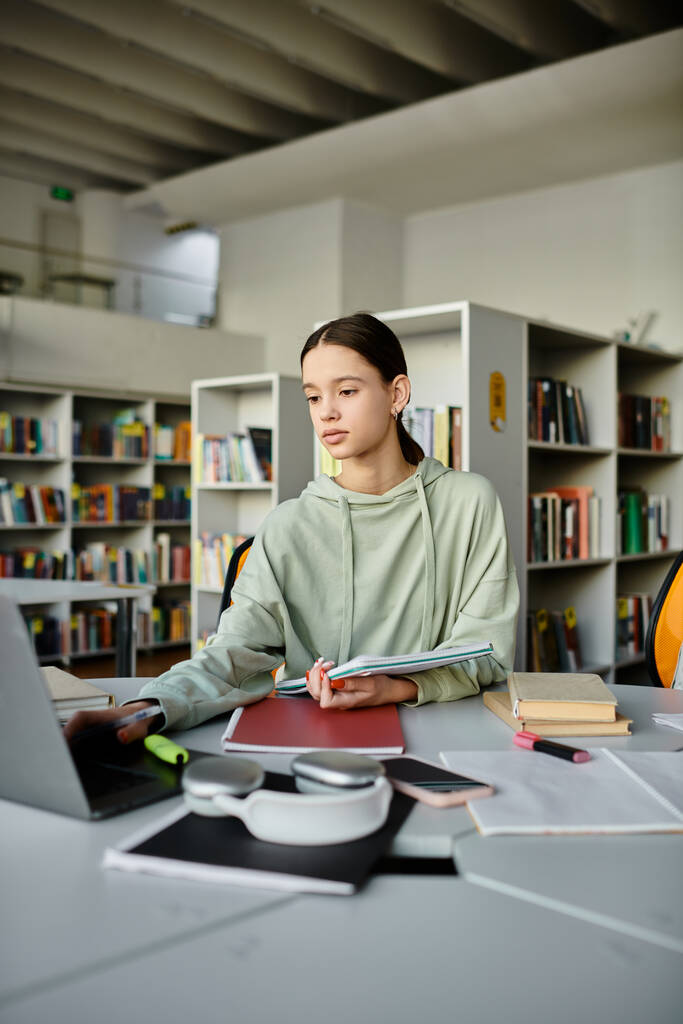 Ένα έφηβο κορίτσι κάθεται σε ένα γραφείο σε μια βιβλιοθήκη, εστιάζοντας έντονα στο laptop της, ενώ κάνει την εργασία μετά το σχολείο. - Φωτογραφία, εικόνα