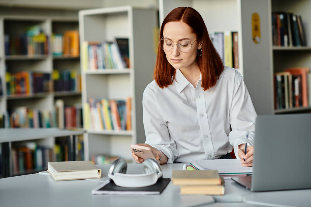 Egy vörös hajú tanárnő online órákat tart, egy íróasztalnál ül egy könyvtárban egy laptoppal.. - Fotó, kép
