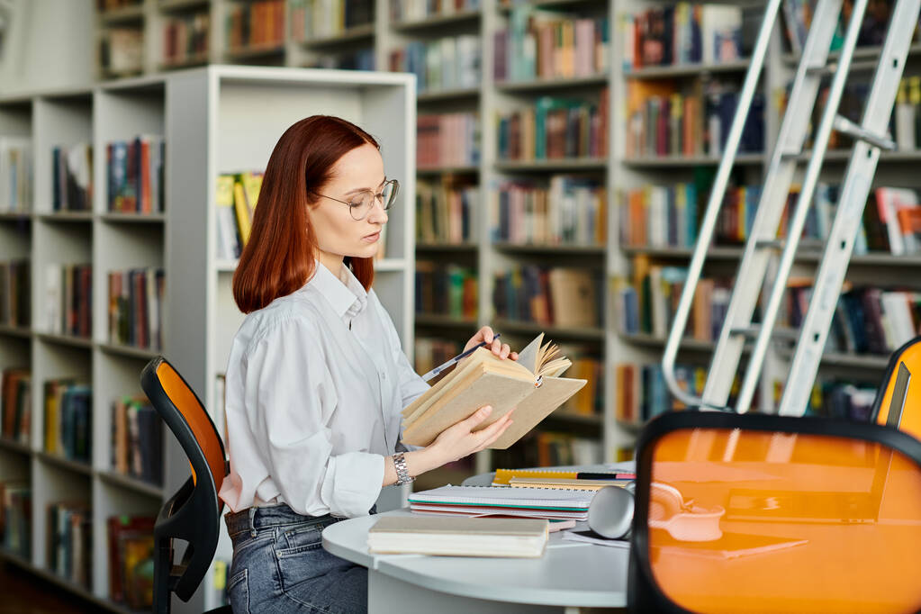 Una donna dai capelli rossi siede a un tavolo in una biblioteca, assorta nella lettura di un libro. - Foto, immagini