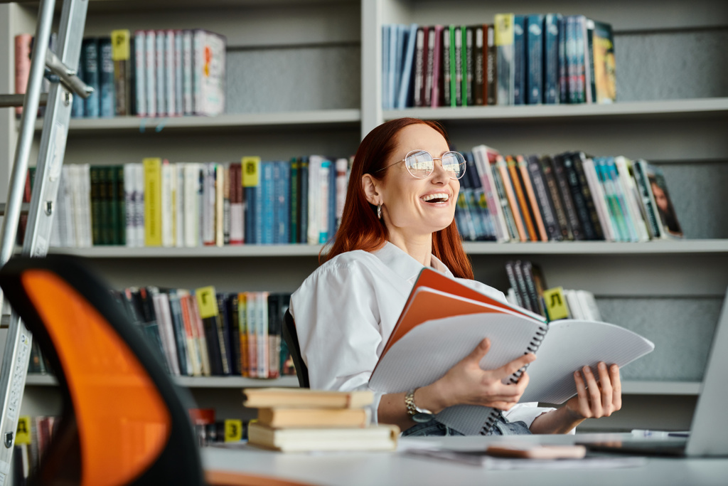 Een roodharige vrouwelijke docent geeft online les, zittend aan een bibliotheekbureau en verdiept in het lezen van een boek na schooltijd.. - Foto, afbeelding