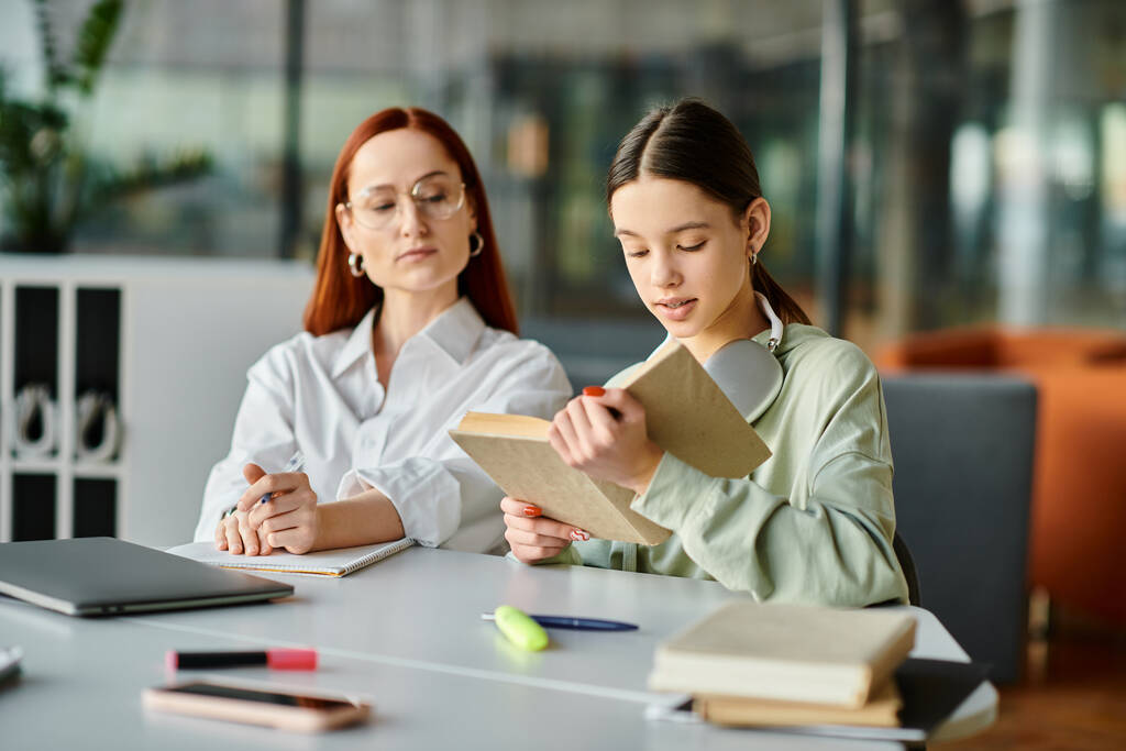 Egy vörös hajú nő tanít egy tinédzser lányt az asztalnál, mindketten egy könyvbe merülve, a közelben laptoppal. Modern oktatás működés közben. - Fotó, kép