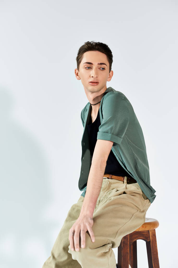若い男は自信を持ってスタイリッシュな緑のシャツとタンパンツのスツールの上に座って灰色のスタジオの背景. - 写真・画像