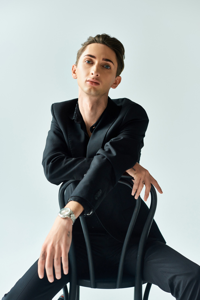 Een jonge homo straalt zelfvertrouwen en trots uit terwijl hij in een zwart pak op een stoel in een studio zit tegen een grijze achtergrond. - Foto, afbeelding