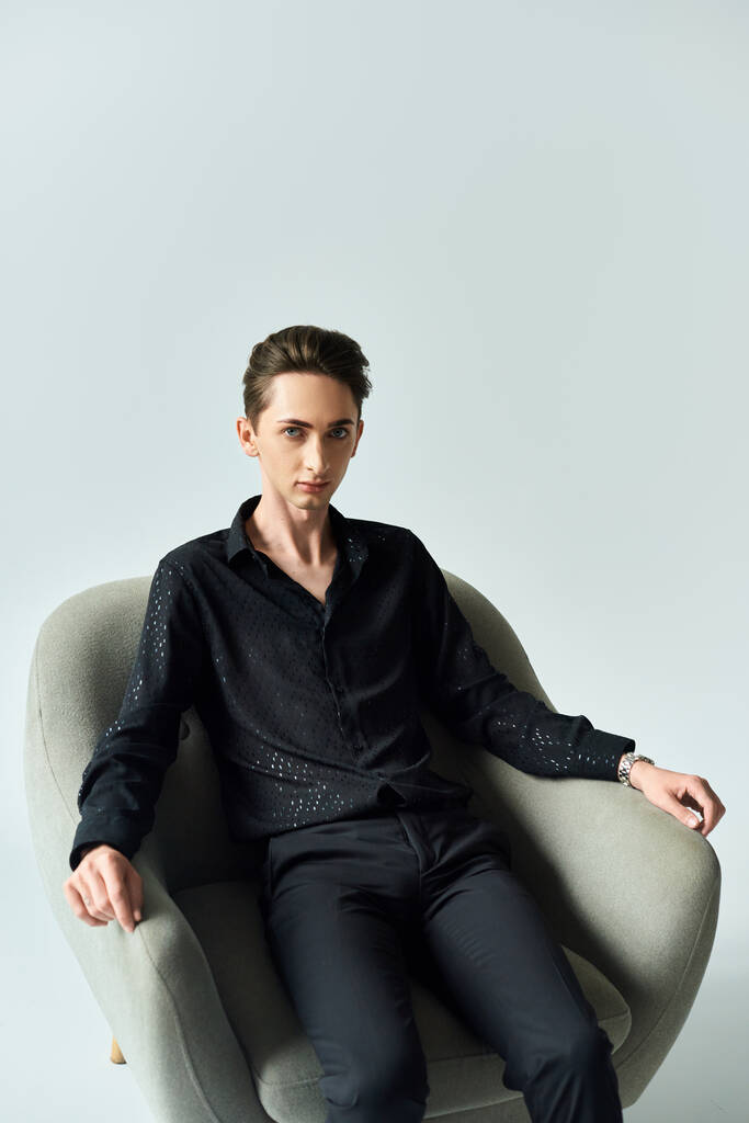 Un giovane uomo queer posa fiducioso in una camicia nera mentre è seduto su una sedia su uno sfondo grigio studio. - Foto, immagini