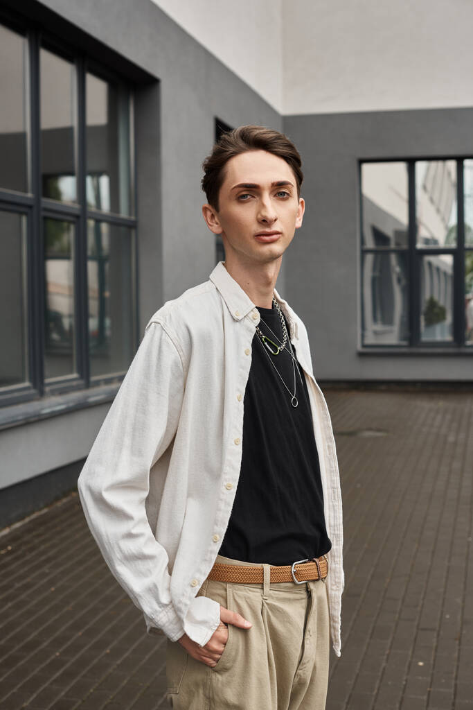 Een jonge homo straalt vertrouwen uit in zijn witte shirt en bruine broek, omarmt zijn unieke stijl met een vleugje LGBTQ trots. - Foto, afbeelding
