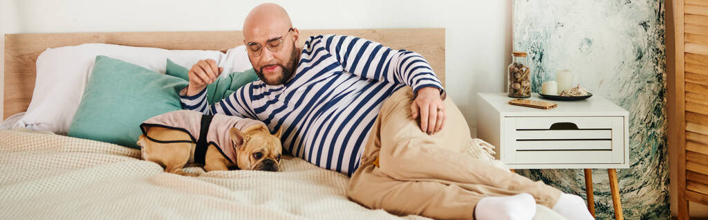 Jóképű szemüveges férfi békésen fekszik a francia bulldogja mellett egy kényelmes ágyon.. - Fotó, kép