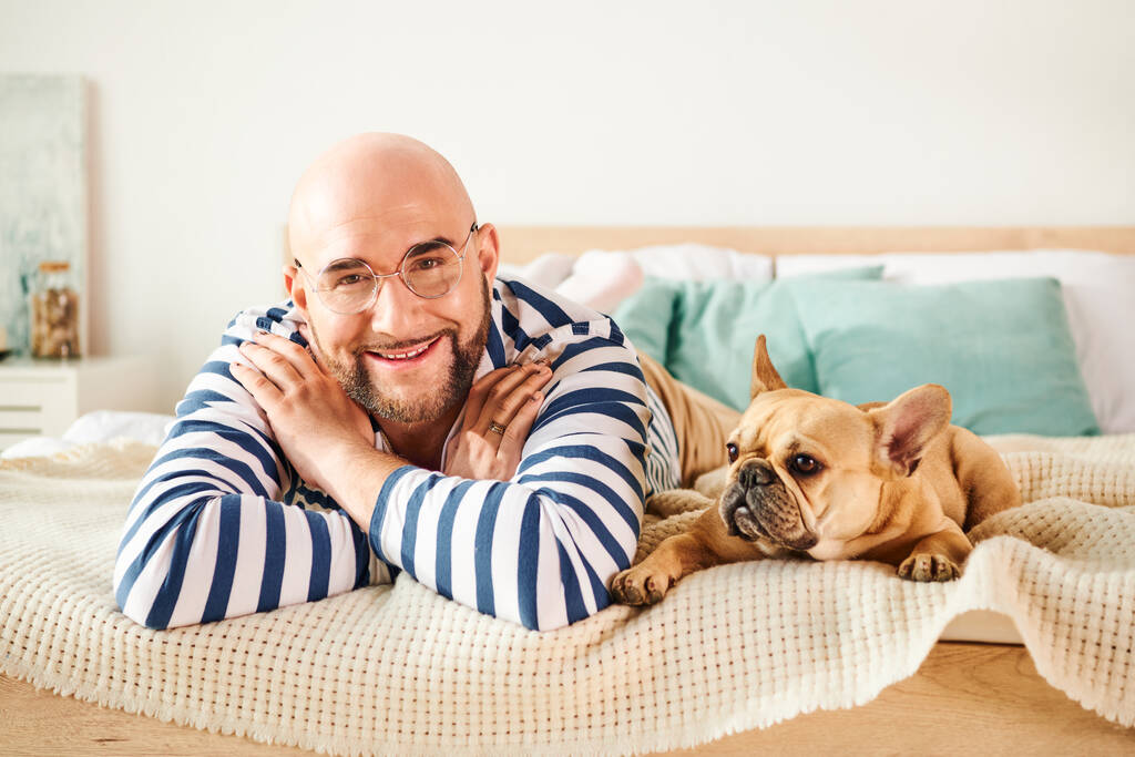 Красивый мужчина в очках расслабляется на кровати рядом со своим верным французским бульдогом. - Фото, изображение