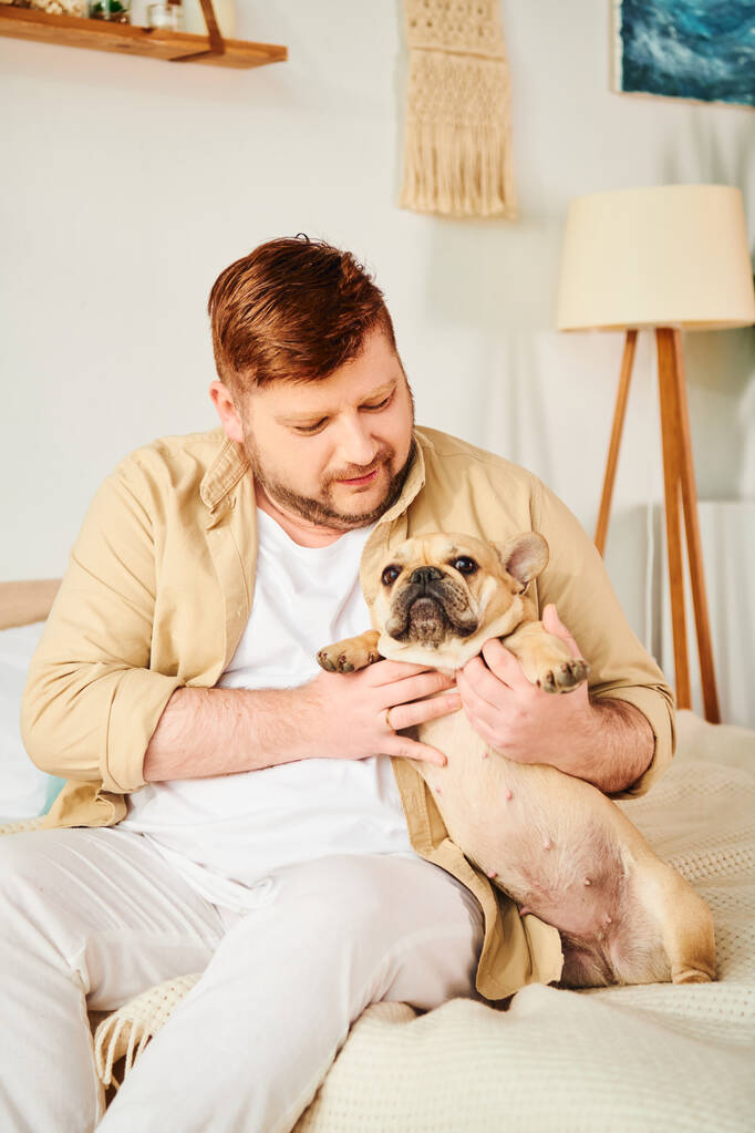Ein Mann sitzt auf einem Bett und hält liebevoll eine kleine französische Bulldogge. - Foto, Bild