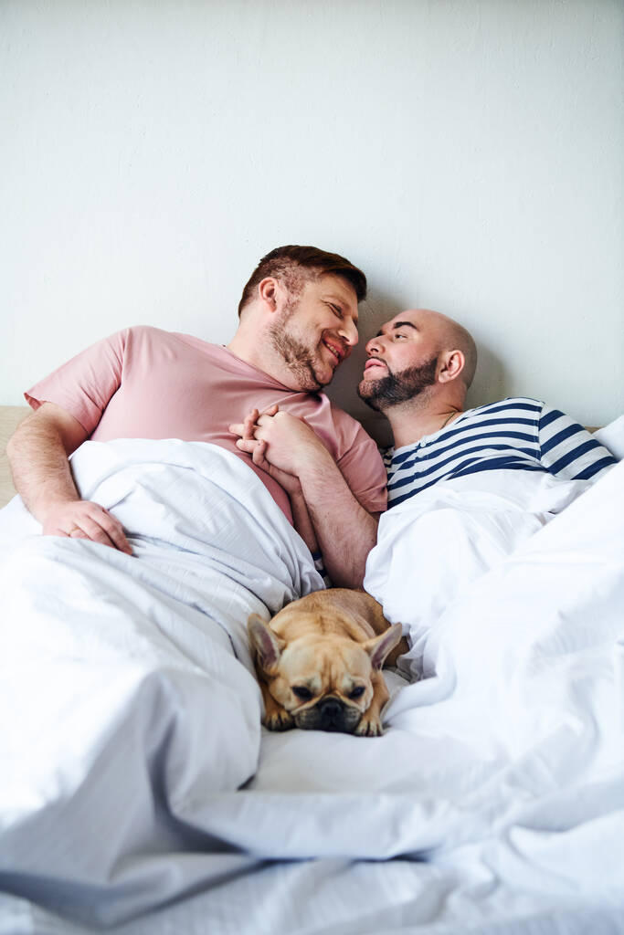 Пара чоловіків, що сидять у ліжку зі своїм французьким бульдогом. - Фото, зображення