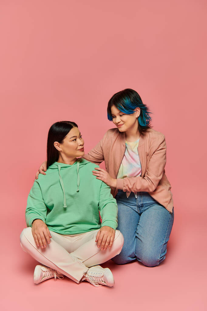 Μια Ασιάτισσα μητέρα και η έφηβη κόρη της κάθονται η μία δίπλα στην άλλη σε ένα στούντιο, φορώντας και οι δύο καθημερινά ρούχα, σε ροζ φόντο.. - Φωτογραφία, εικόνα