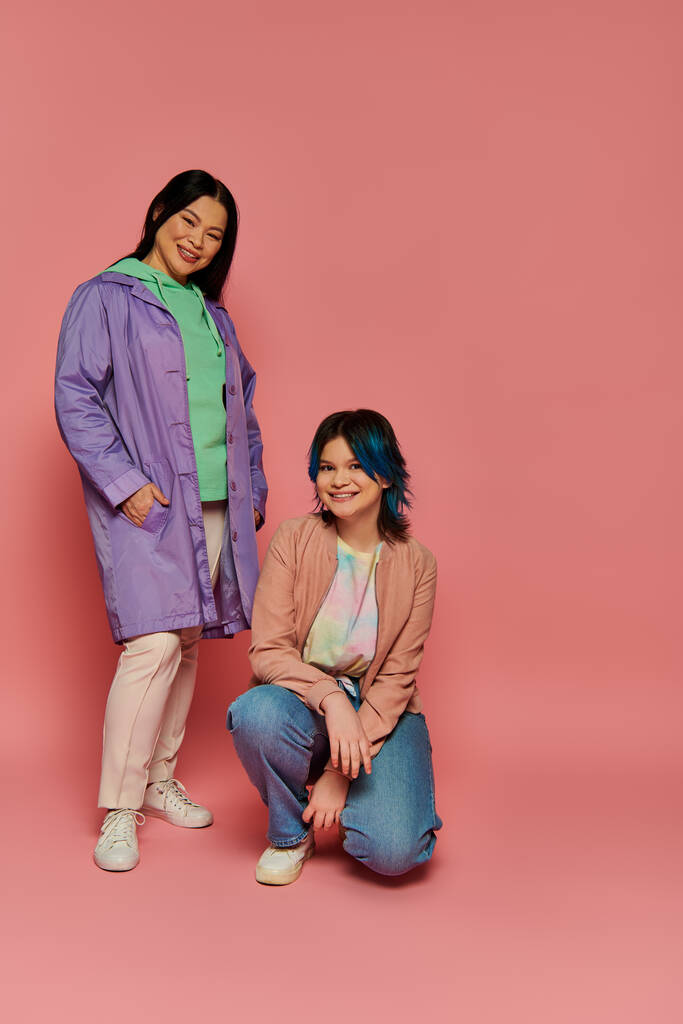 Aziatische moeder en haar tienerdochter poseren vol vertrouwen in de voorkant van een roze achtergrond in een stijlvolle studio setting. - Foto, afbeelding