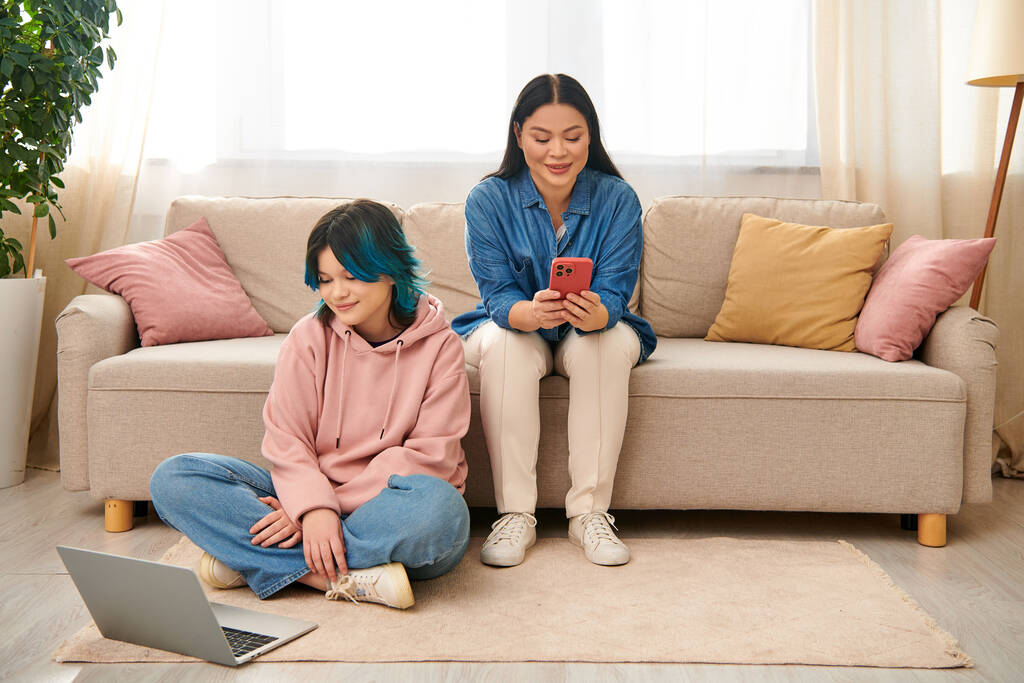 Мать-азиатка и ее дочь-подросток, одетые в повседневную одежду, сидят на диване и сосредоточены на экране мобильного телефона. - Фото, изображение