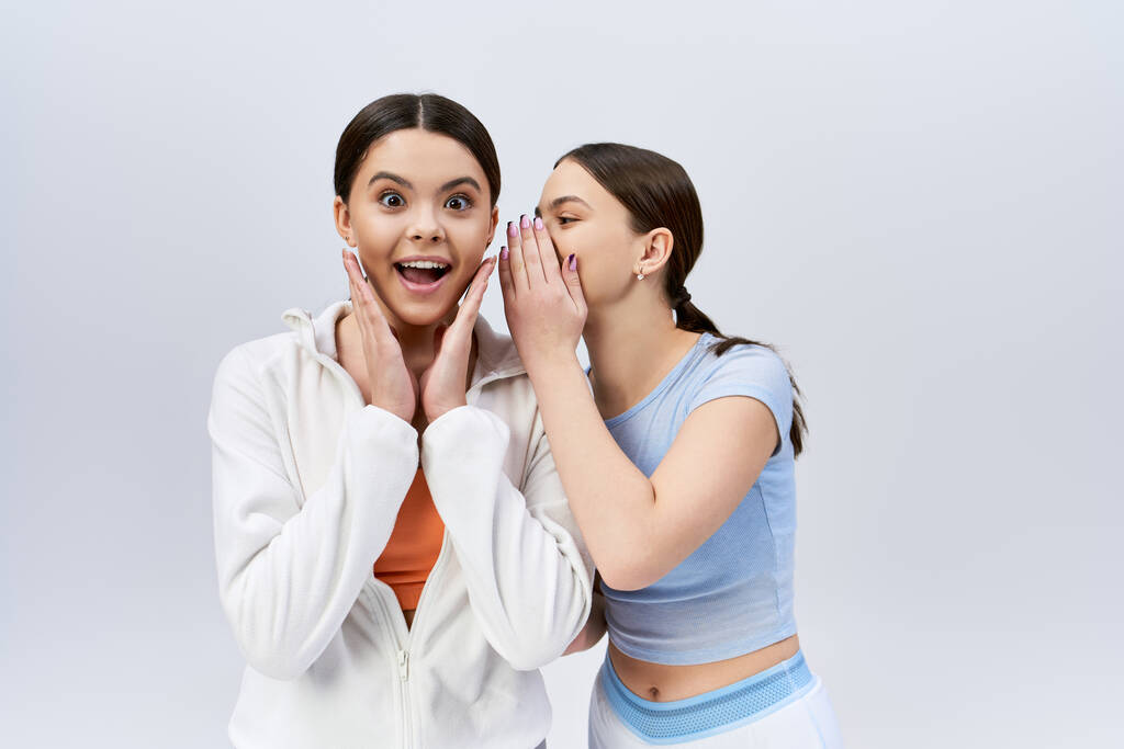 Twee mooie, brunette tienermeisjes in sportieve kleding staan zij aan zij, handen op gezichten, in een studio op een grijze achtergrond. - Foto, afbeelding