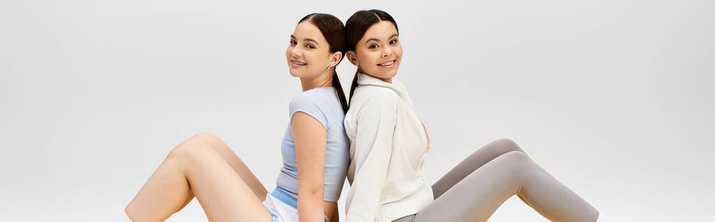 Two pretty, young women, brunette and dressed in sportive attire, sitting closely together in a studio against a grey background. - Photo, Image